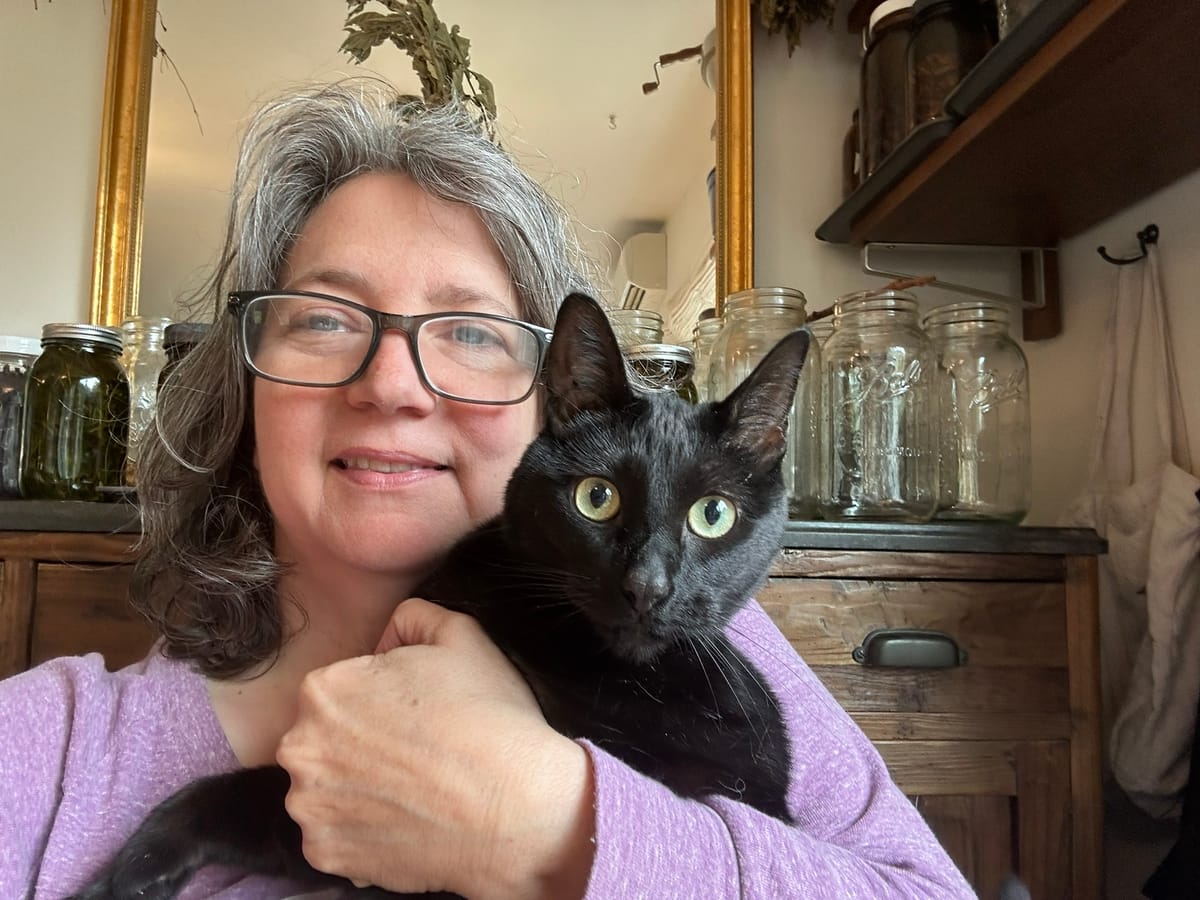 A photo of me--a fierce middle-aged white woman with glasses and gray hair--holding an even fiercer black cat starring straight at the camera 