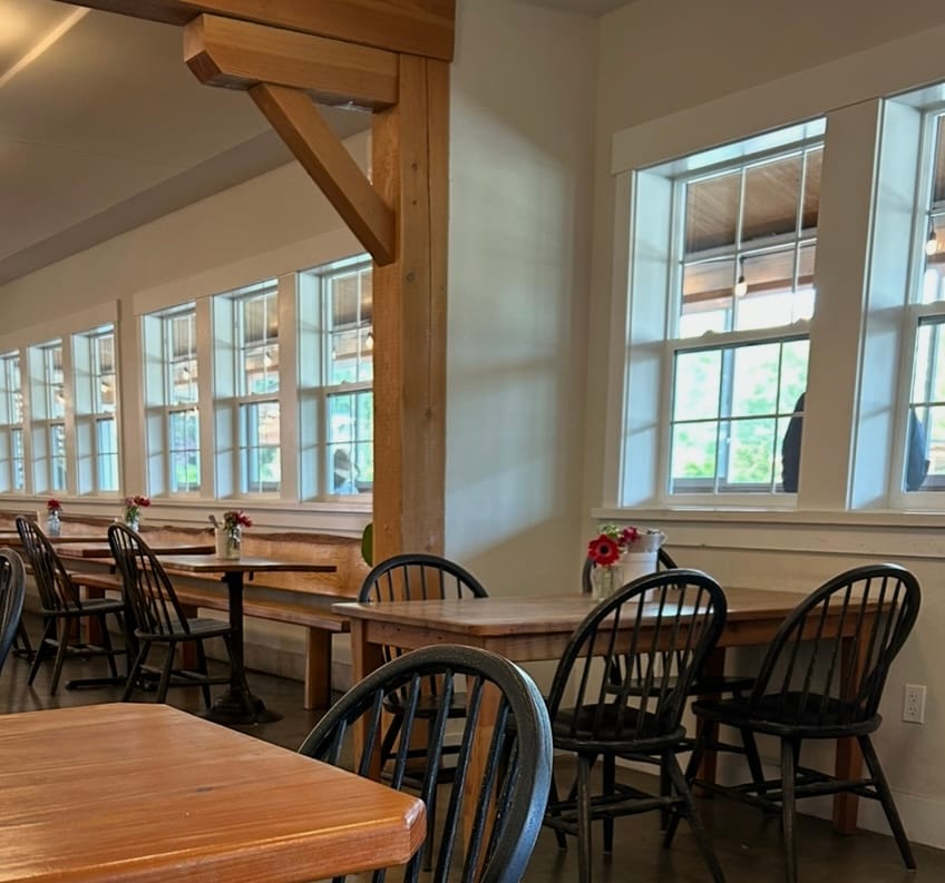 Photo of an empty diner with wooden tables and chairs and a white windows and a person whose arms you can see but whose face and center body you can't see standing outside