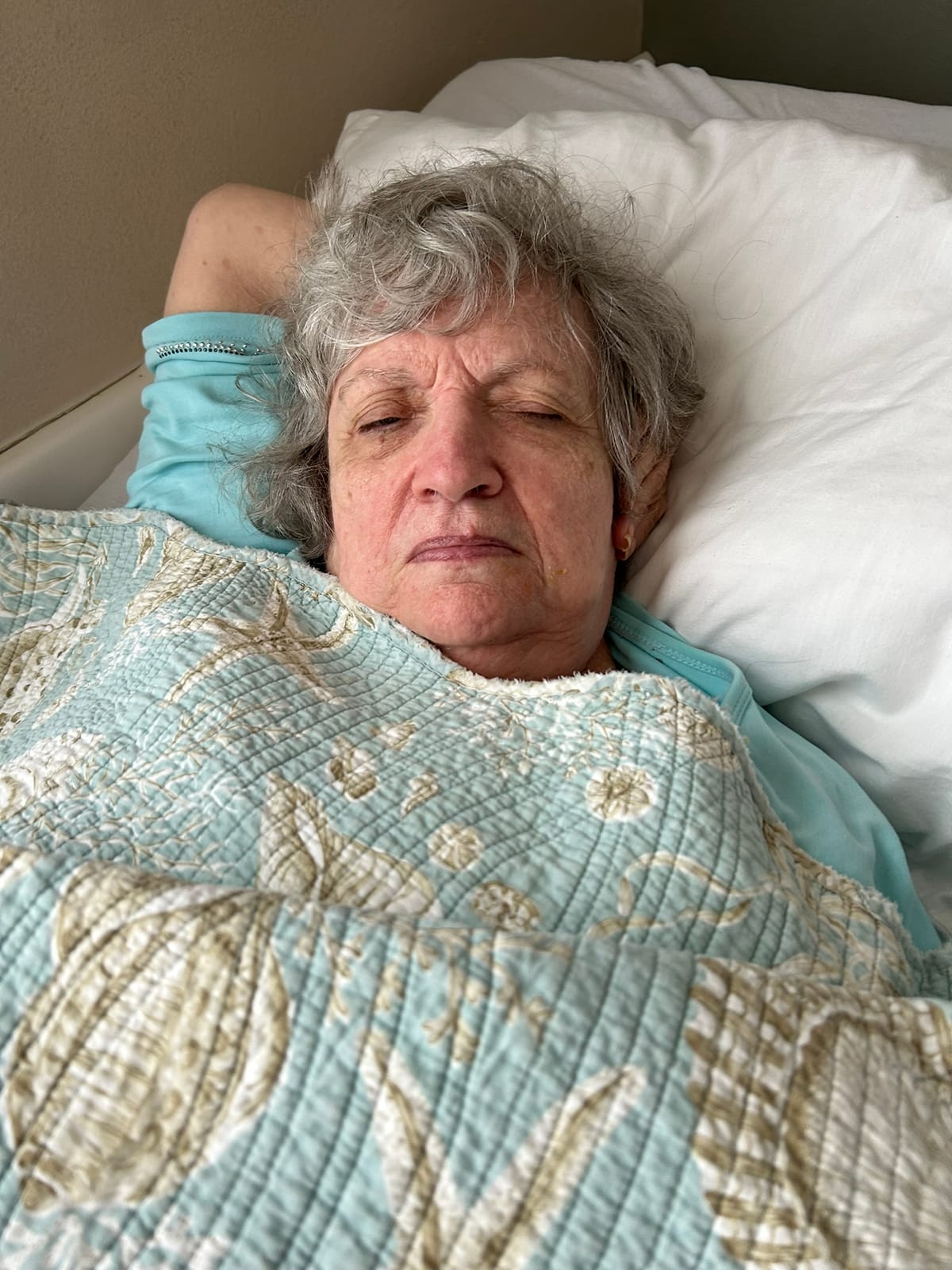 A gray-haired elder woman with her eyes almost closed lays in bed under a light blue comforter with seashells on it.