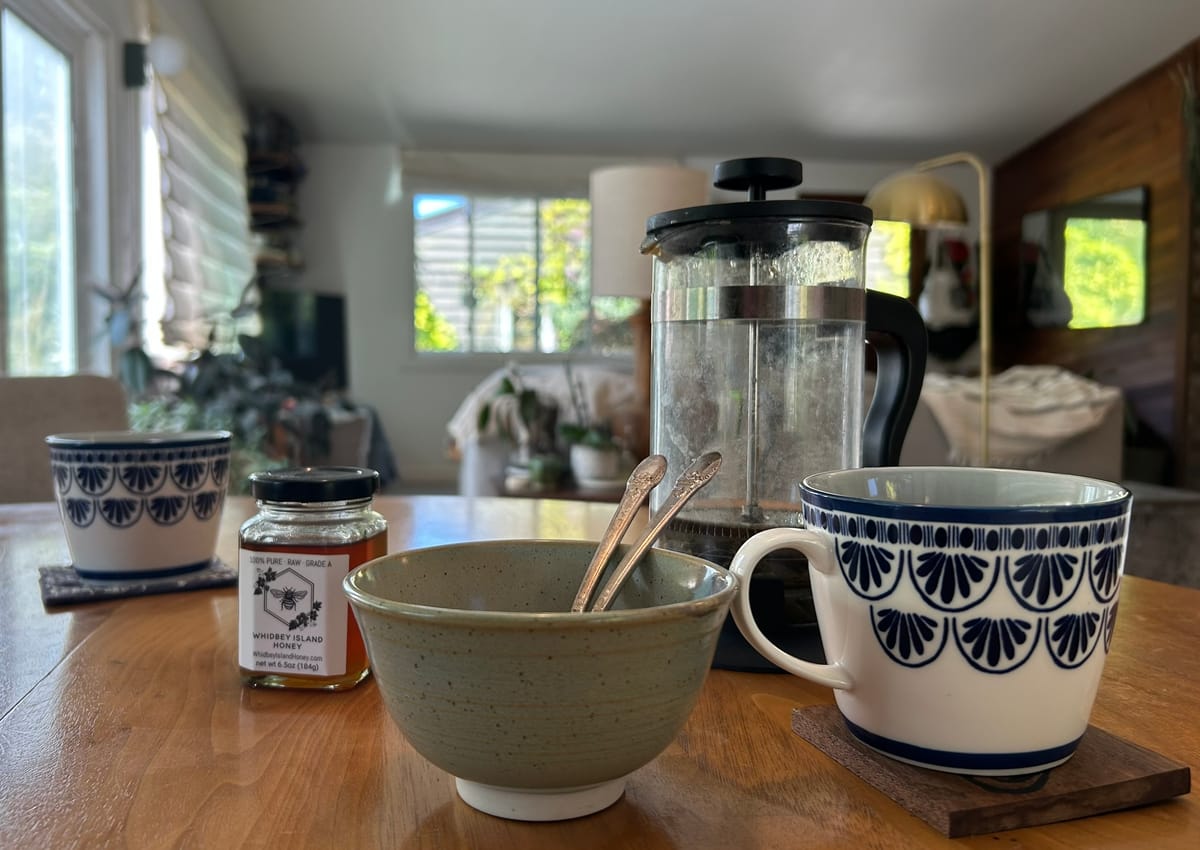 Close-up photo of two blue and white teacups with a jar of honey, a bowl holding spoons, and a French press on a wood table.