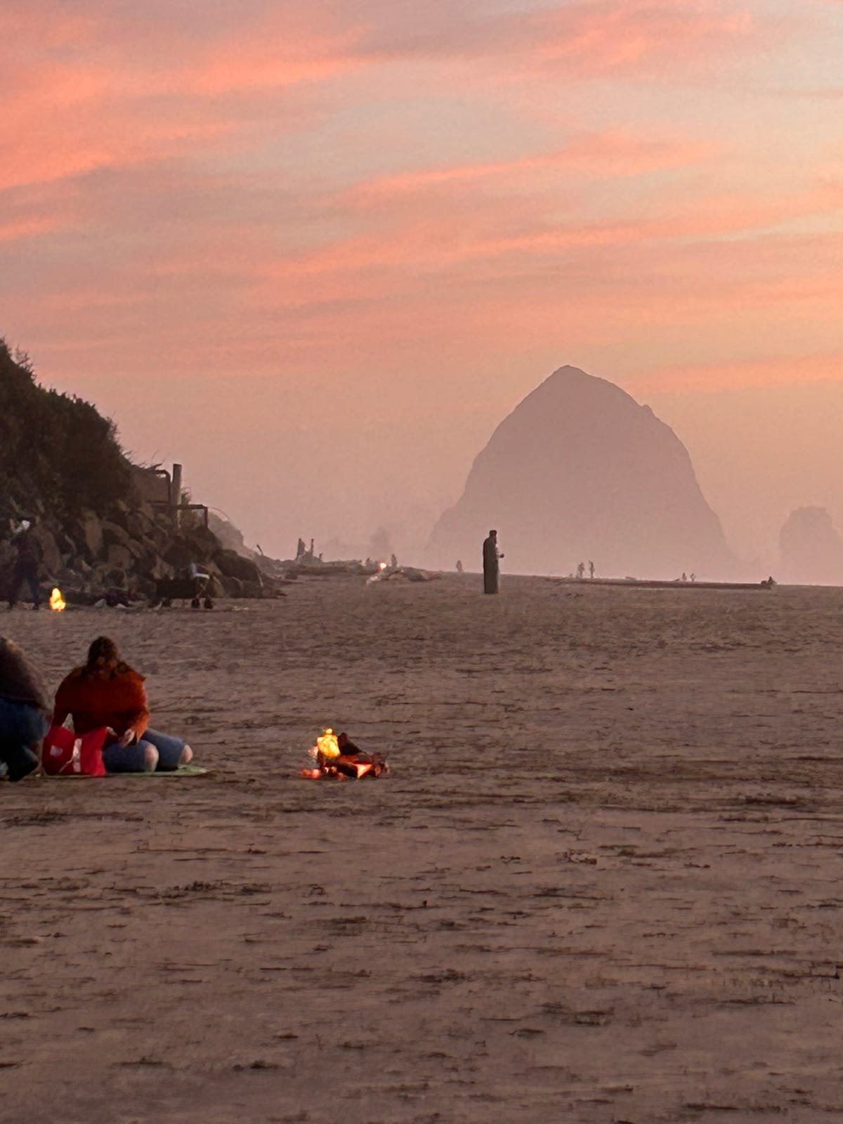 A photo of a beach at sunset with people in the distance sitting around fires and walking on the beach 