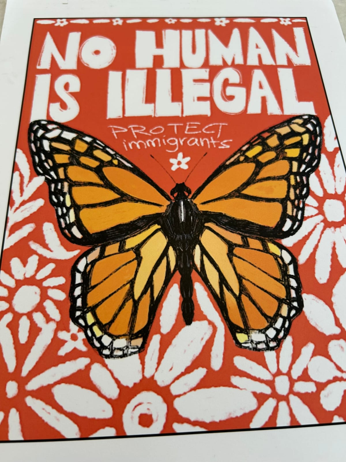 A bright orange poster with white flowers and a monarch butterfly on it that says "No Human is Illegal. Protect immigrants."