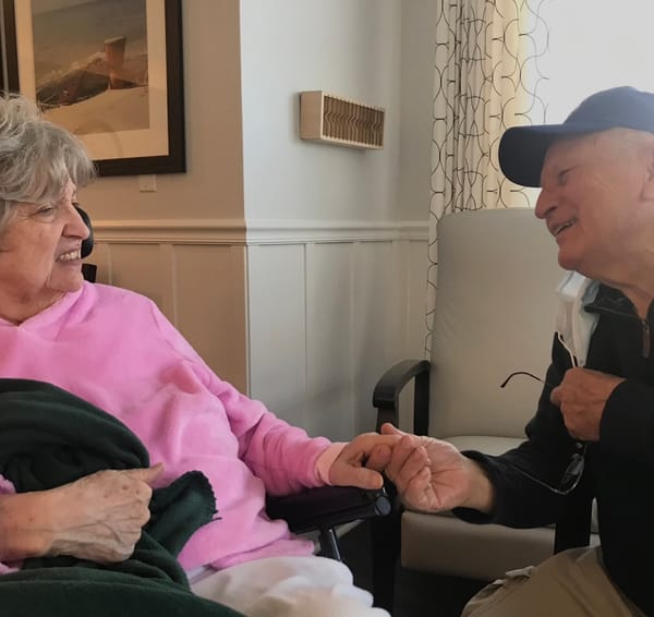 Two elders with gray hair-- a woman in a pink shirt and a man in a blue coat and hat--hold hands and look lovingly at each other. 