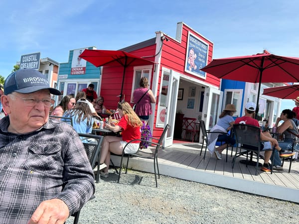 Old man in a gray flannel shirt and dark blue Birdsview Brewery baseball cap sits in front of a red hotdog stand with lots of patrons sitting on the deck behind him on a sunny summer day