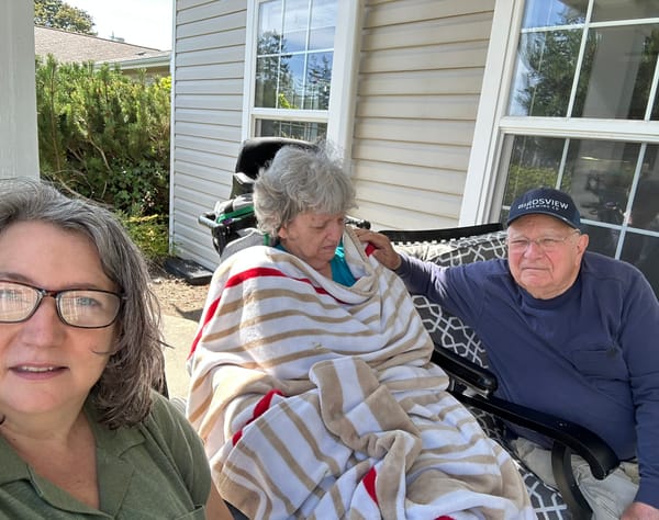 One middle-aged white woman in a moss green shirt and her elder parents, Dad in blue, Mom wrapped in a stripped blanked, outside a tan colored memory care home