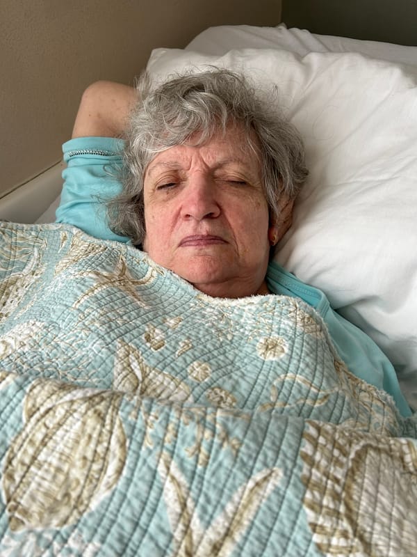 A gray-haired elder woman with her eyes almost closed lays in bed under a light blue comforter with seashells on it.