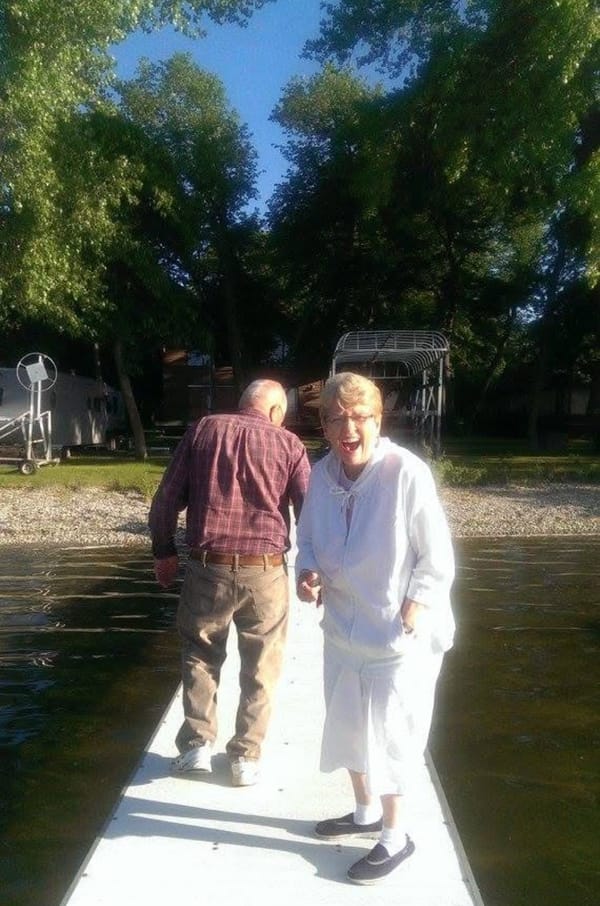 An old woman in white, laughing at the camera, standing on a dock out over the water, beside an old man in a red shirt, walking away. Who she'd considered pushing in the lake.