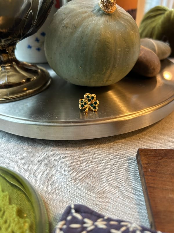 A photo of a gold clover-shaped earring with green emeralds in the leaves, surrounded by two colored glass vases, a green gourd, and blue and brown coasters