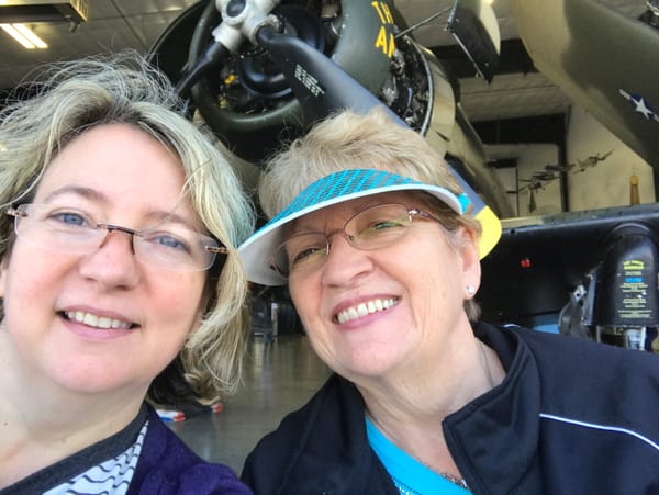 Two white women--one middle aged and one older--with blond-ish hair, glasses, and smiles take a selfie in the wind in front of an old airplane